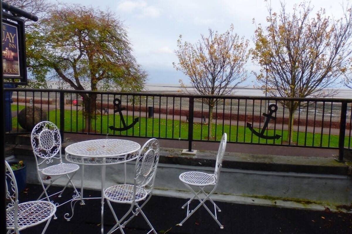 A patio with white metal chairs and tables, a black metal fence, and a view of a grassy area with trees and a body of water.