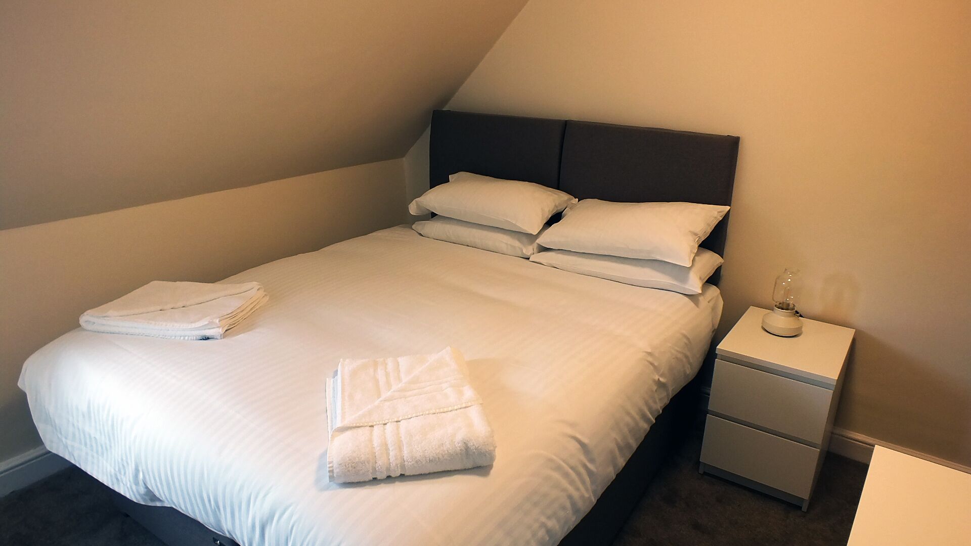 Una cama bien hecha con sábanas y almohadas blancas, una toalla doblada sobre la cama y una mesita de noche con una lámpara.