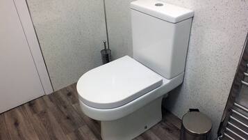 A modern toilet with a white tank and seat, a metal bidet sprayer, and a small trash bin.