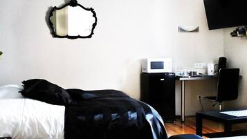 A hotel room with a bed, a desk with a computer, a microwave, a chair, and a flat-screen TV mounted on the wall.
