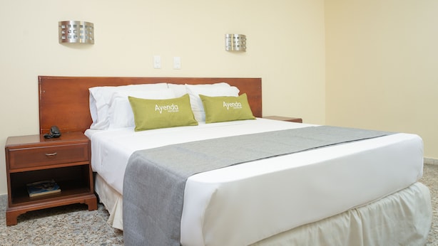 Una habitación de hotel con cabecera de madera, dos almohadas con la marca 'Ayenda Hotels', una mesita de noche con un libro y una manta gris.