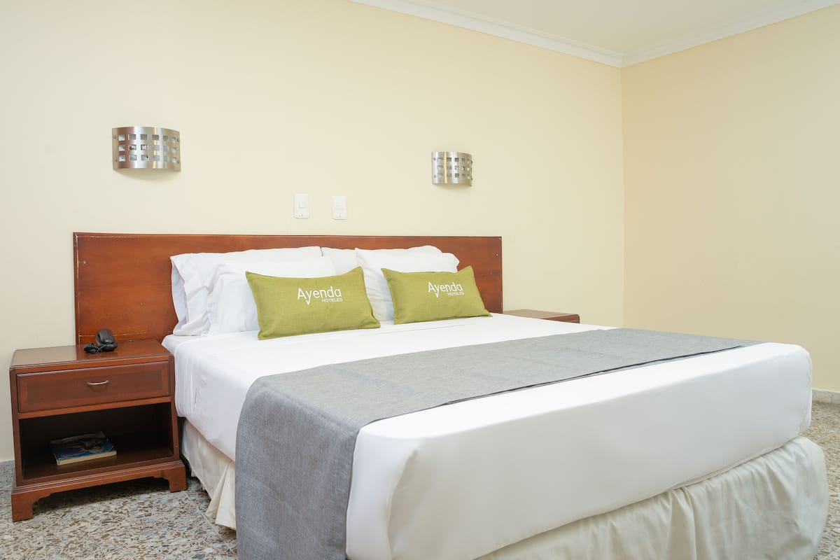 Una habitación de hotel con cabecera de madera, dos almohadas con la marca 'Ayenda Hotels', una mesita de noche con un libro y una manta gris.