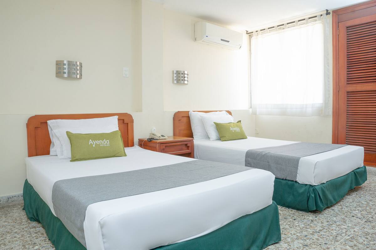 Habitación de hotel con dos camas, ventana con persianas, aire acondicionado y puerta de madera.