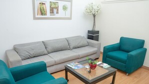 Una sala de estar con un sofá gris, sillones azul verdosos, una mesa de centro de vidrio con revistas, una planta en maceta y un cuadro en la pared.