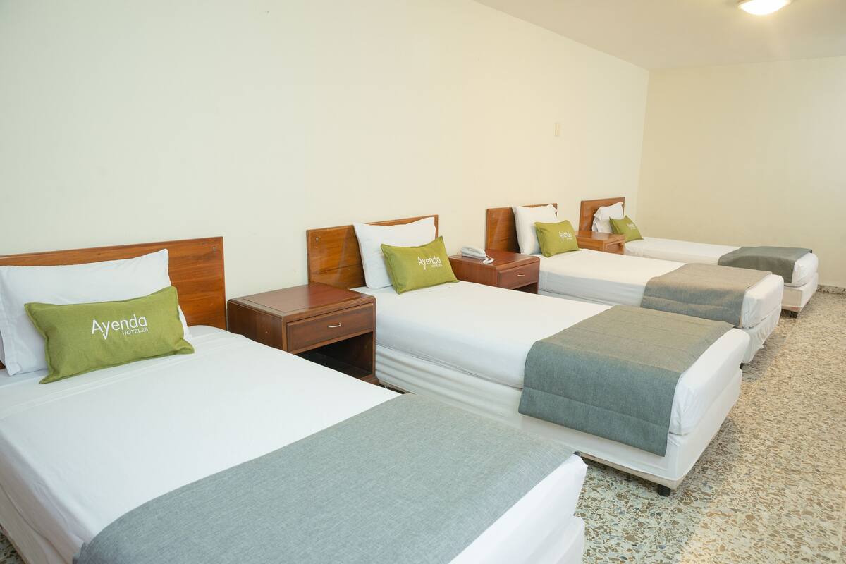 Habitación de hotel con cuatro camas individuales, cada una con cabecera de madera, ropa de cama blanca y almohadas verdes.