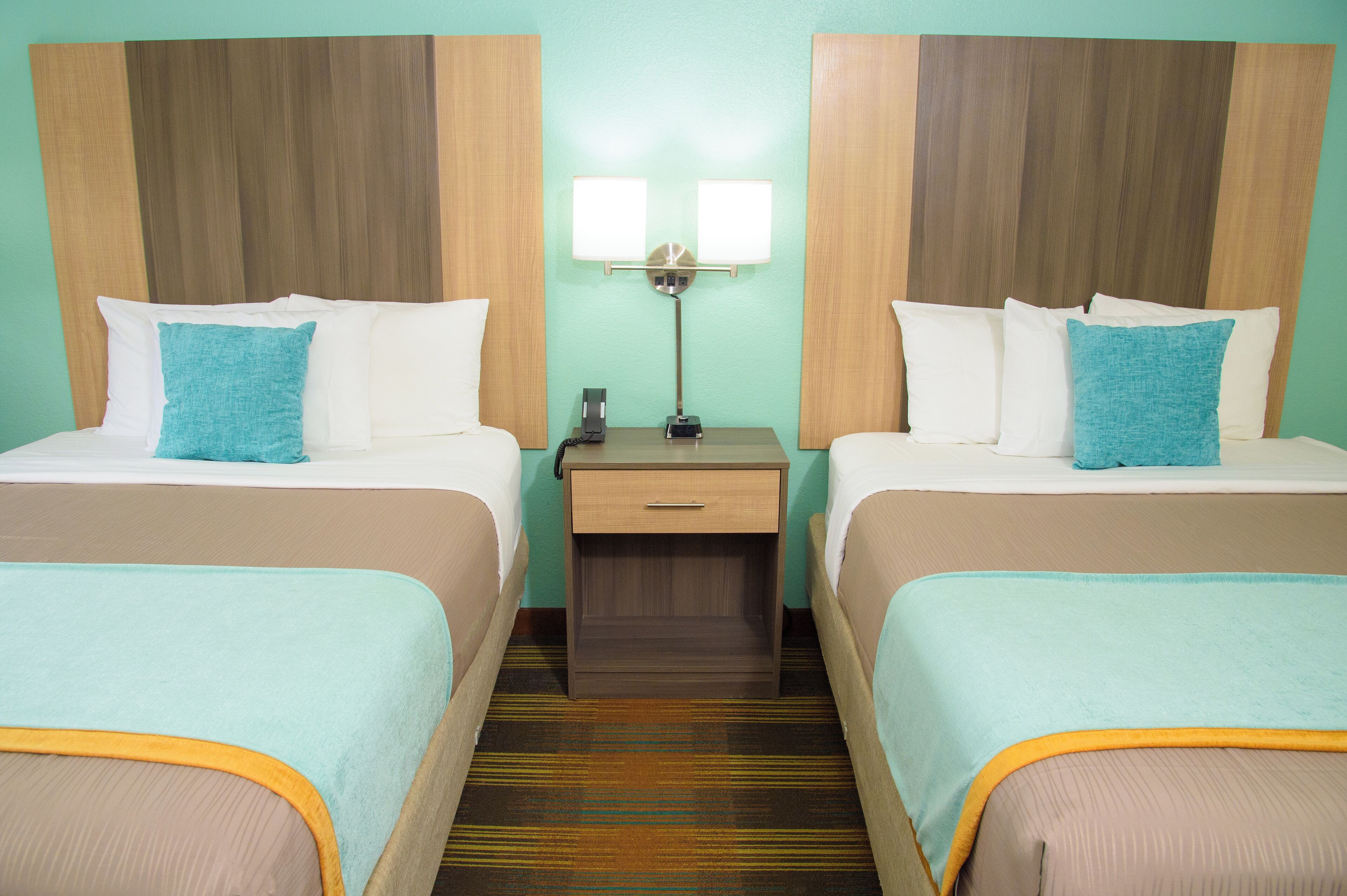 A hotel room with two beds, each with a blue pillow, a nightstand with a lamp, and a wooden headboard.