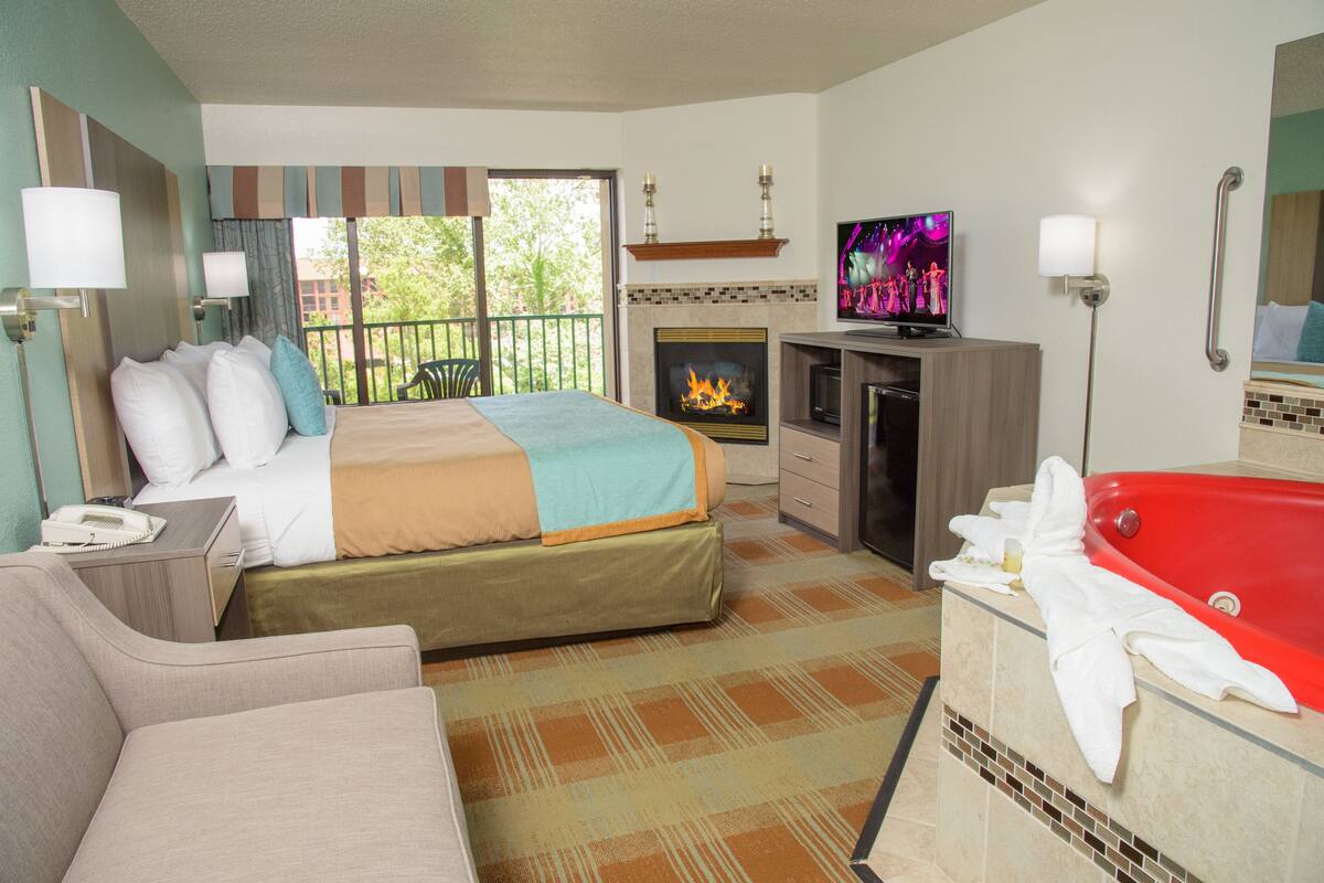 A hotel room with a bed, a TV, a fireplace, a red bathtub, and a balcony with a view of trees.
