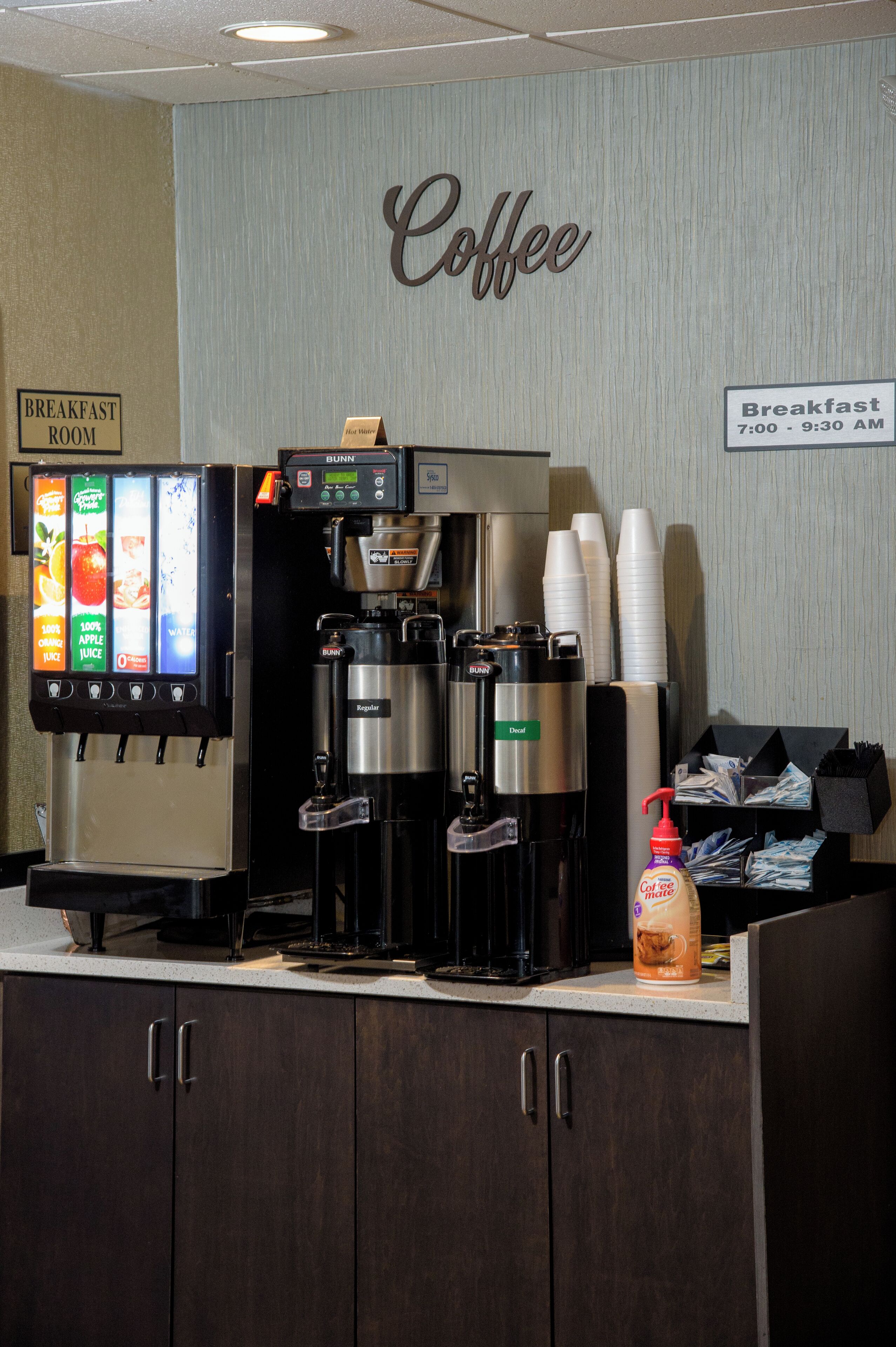 A coffee station with a beverage dispenser, a coffee machine, and a sign indicating breakfast hours.