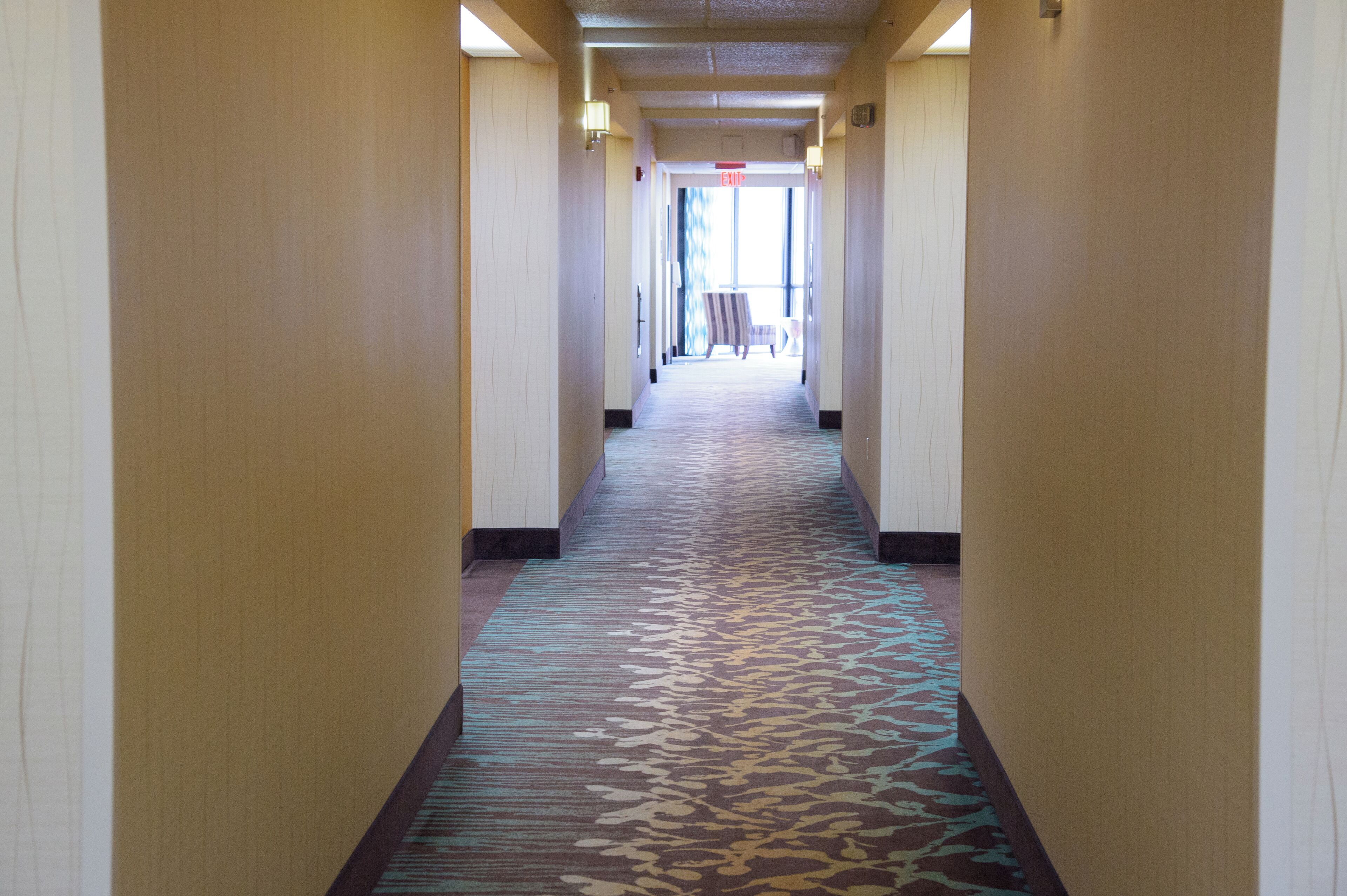 A hotel hallway with a patterned carpet, cream-colored walls, and a door at the end.
