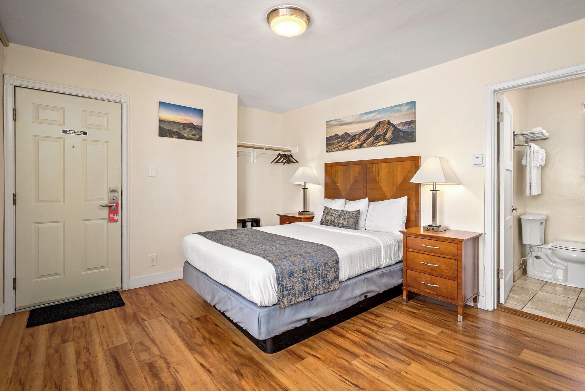 A hotel room with a bed, bedside tables, a wooden headboard, a bathroom with a toilet and sink, and two paintings on the wall.