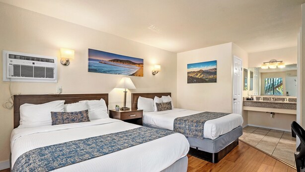 A hotel room with two beds, a nightstand, a lamp, a painting, a mirror, and a view of the ocean. - Sunbeam Motel (San Luis Obispo)