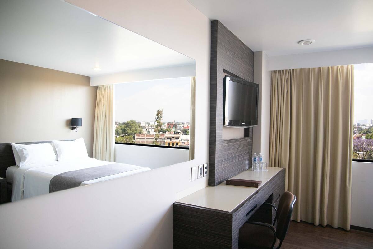 A hotel room with a bed, a desk, a chair, a TV, and a window with curtains.