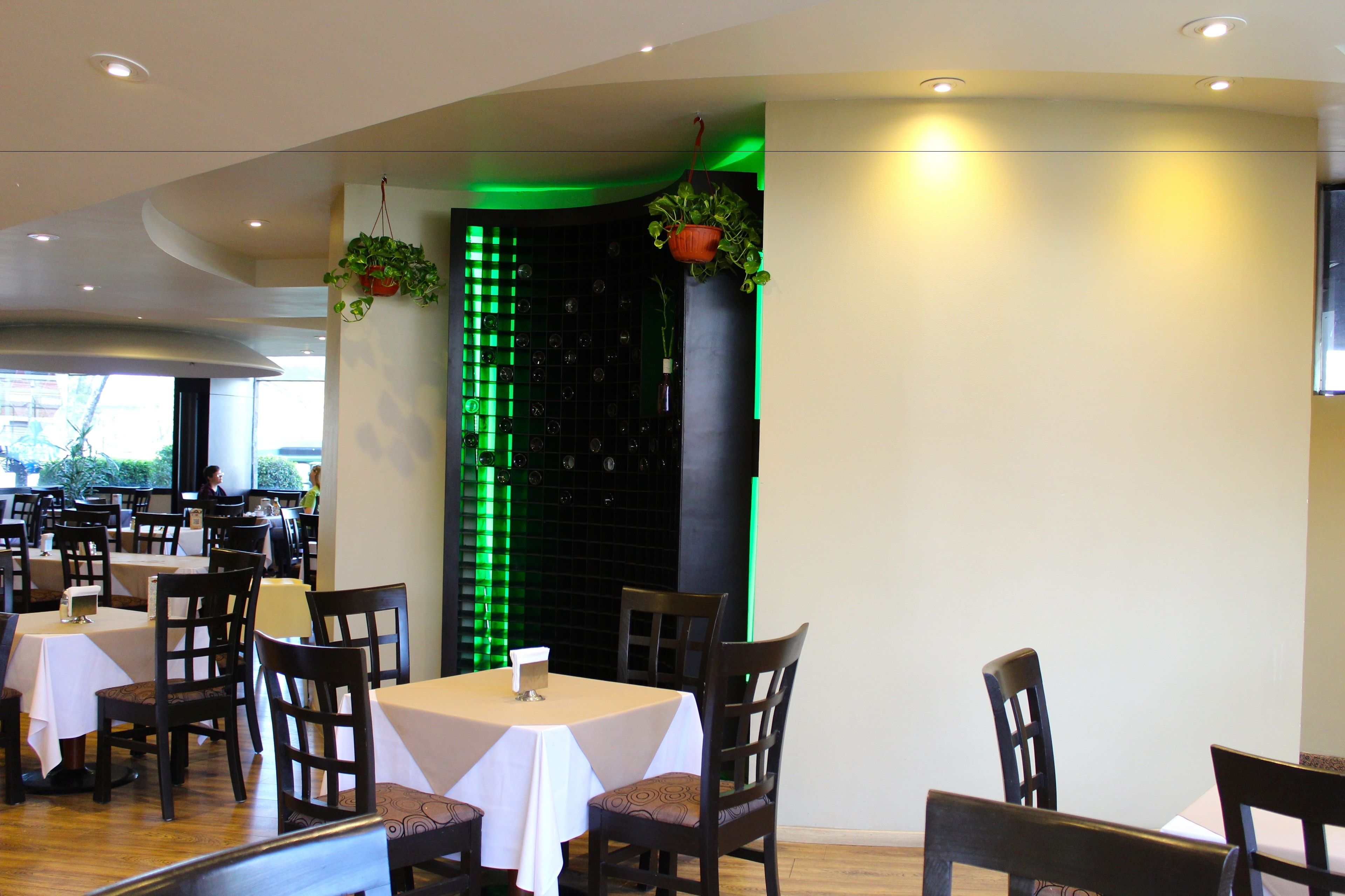A restaurant with multiple tables and chairs, a green illuminated wall, and hanging plants.