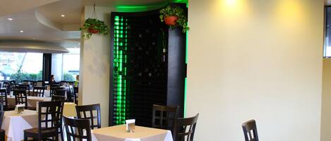 A restaurant with multiple tables and chairs, a green illuminated wall, and hanging plants.