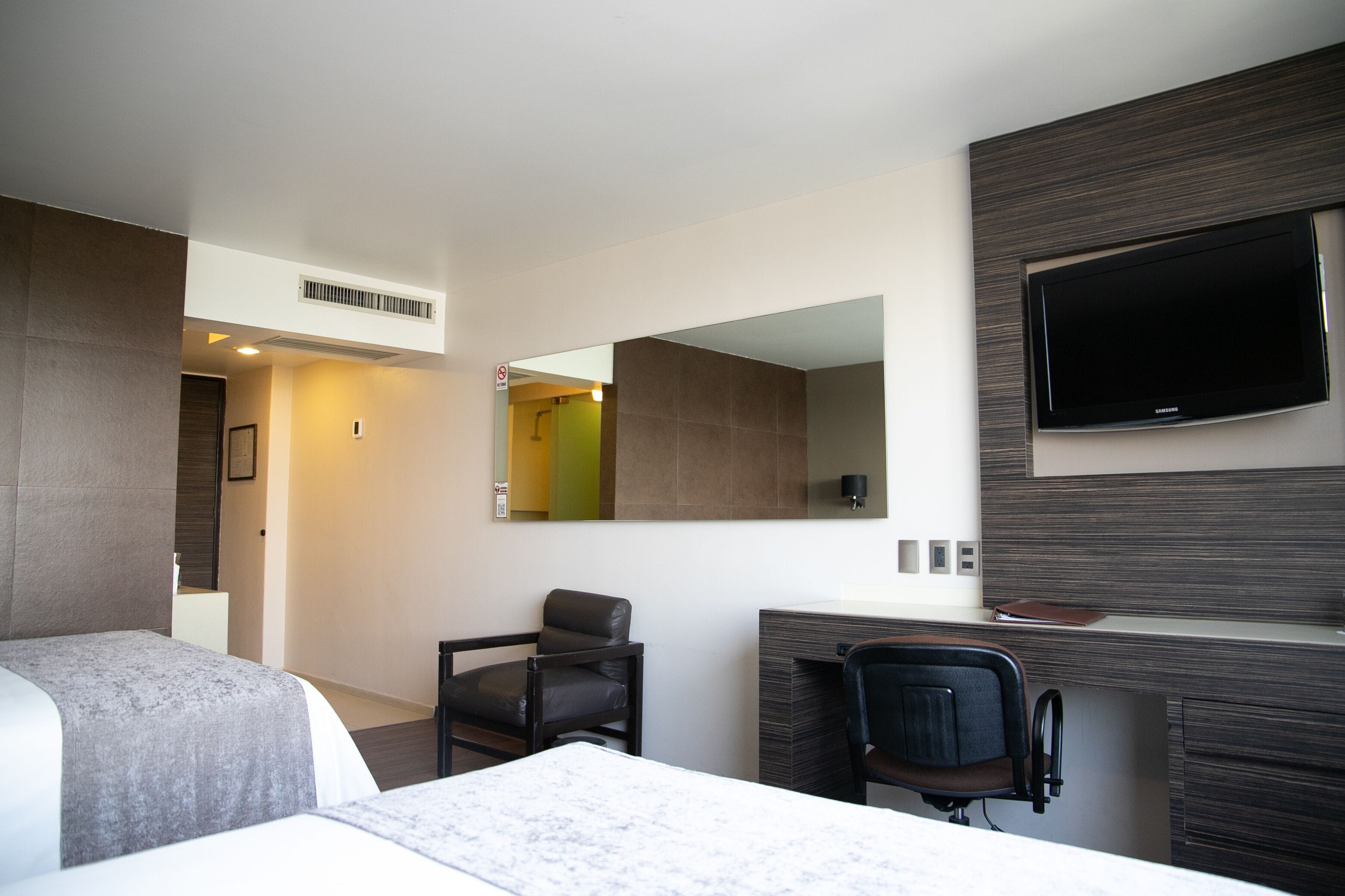 Habitación de hotel con cama, escritorio, silla, televisor y espejo.