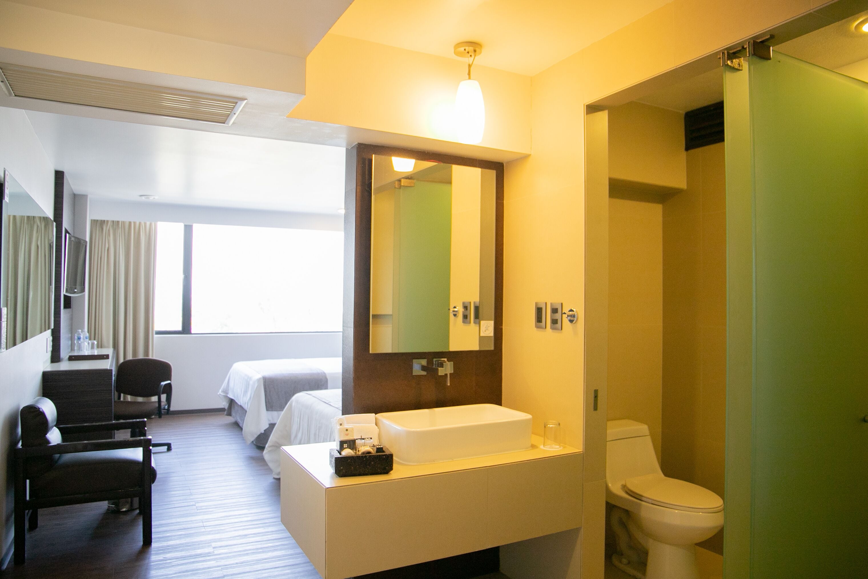 Habitación de hotel con cama, escritorio, silla, espejo grande, lavamanos y retrete.