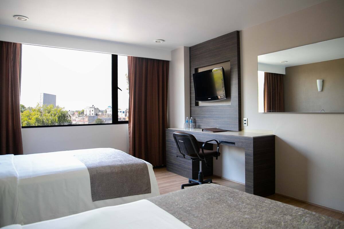 A hotel room with two beds, a desk with a chair, a TV, and a large window with curtains.