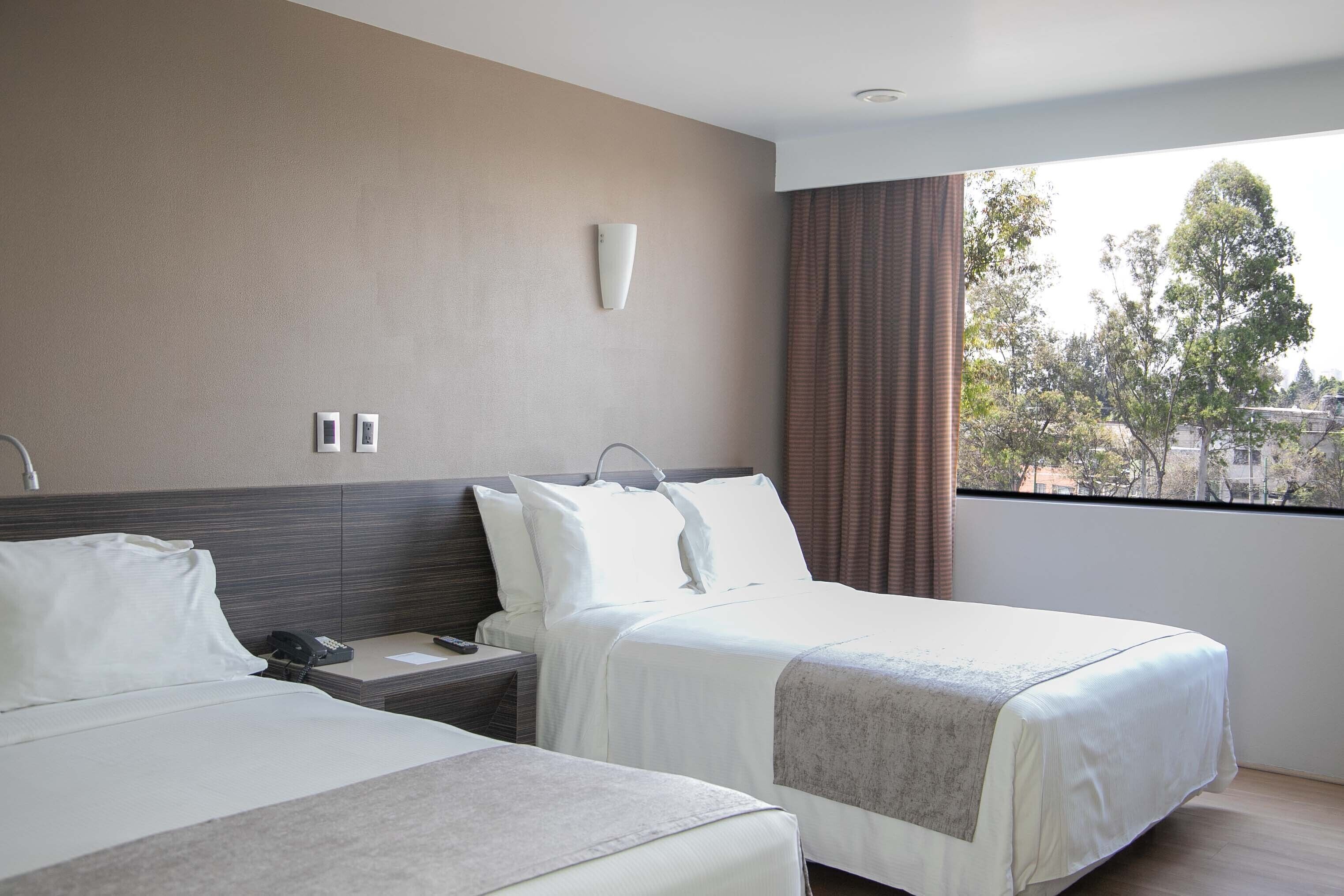 A hotel room with two beds, a bedside table with a phone, a wall-mounted light, and a window with a view of trees and buildings.