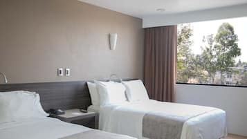 A hotel room with two beds, a bedside table with a phone, a wall-mounted light, and a window with a view of trees and buildings.