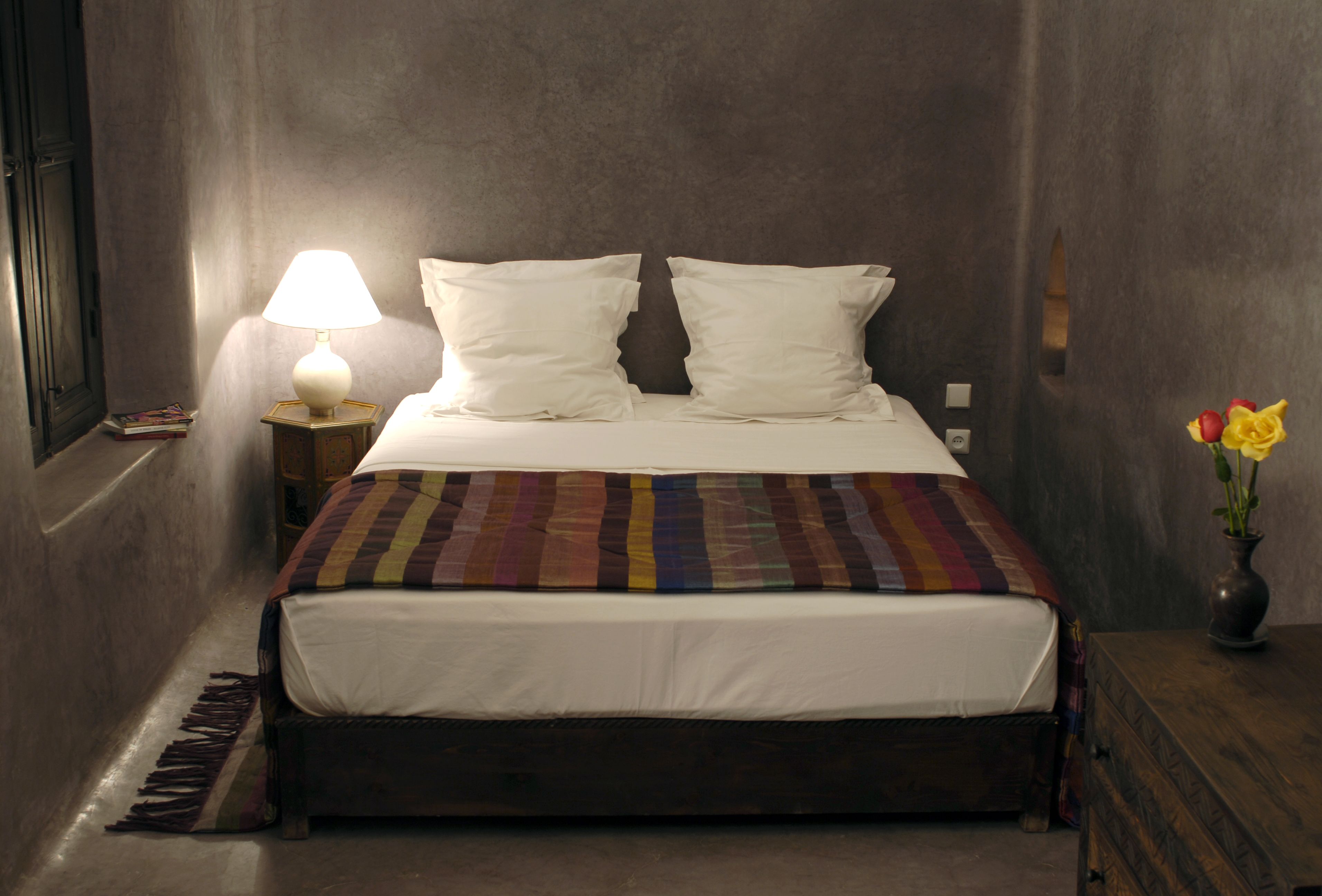 A neatly made bed with a striped blanket, a bedside lamp, and a vase with flowers on a wooden nightstand.
