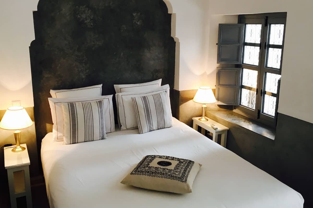 A bed with white bedding and striped pillows, a dark headboard, two bedside lamps, and a window with decorative shutters.