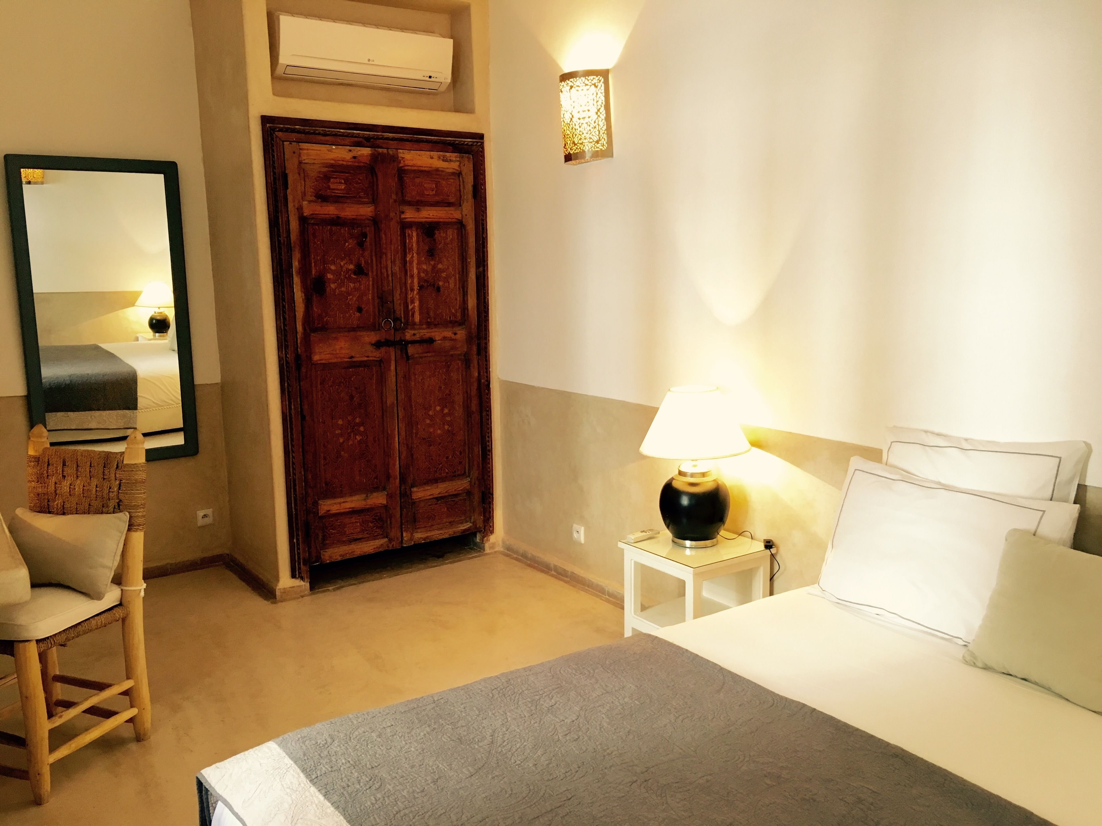 A hotel room with a bed, a chair, a wardrobe, a bedside table with a lamp, and a wall-mounted air conditioner.