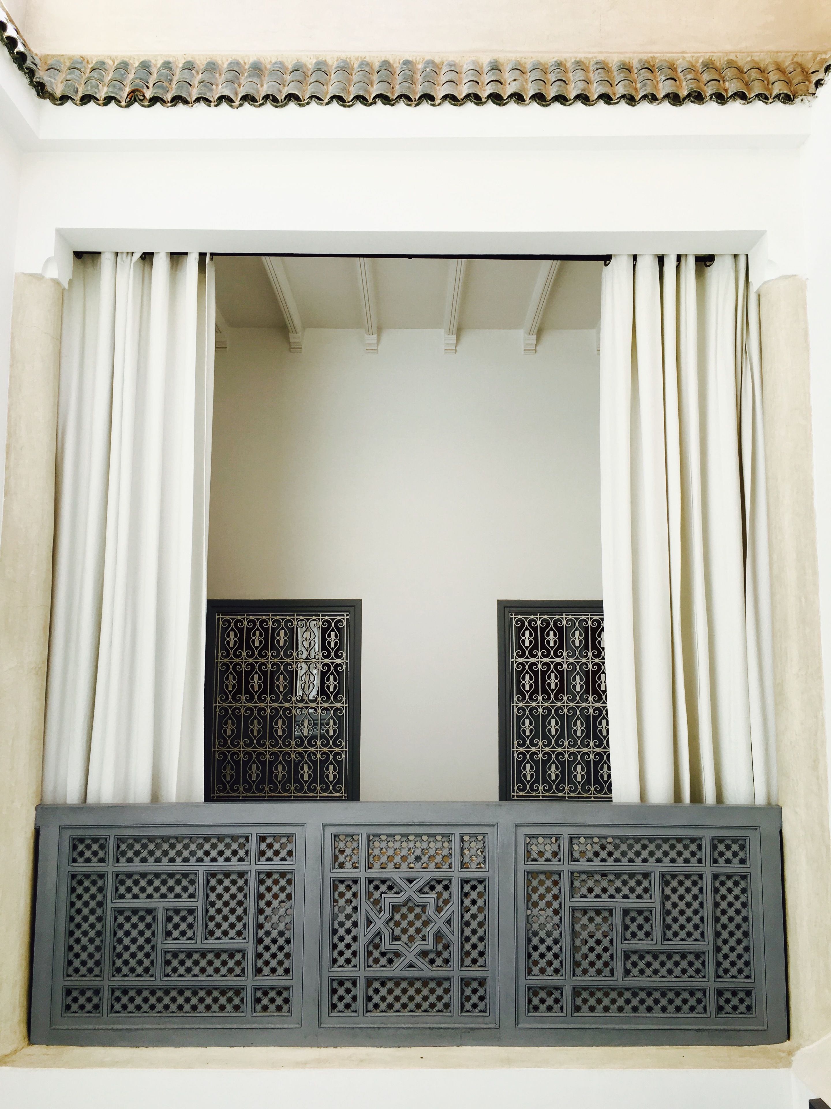 A window with white curtains, a decorative metal grille, and a tiled roof.