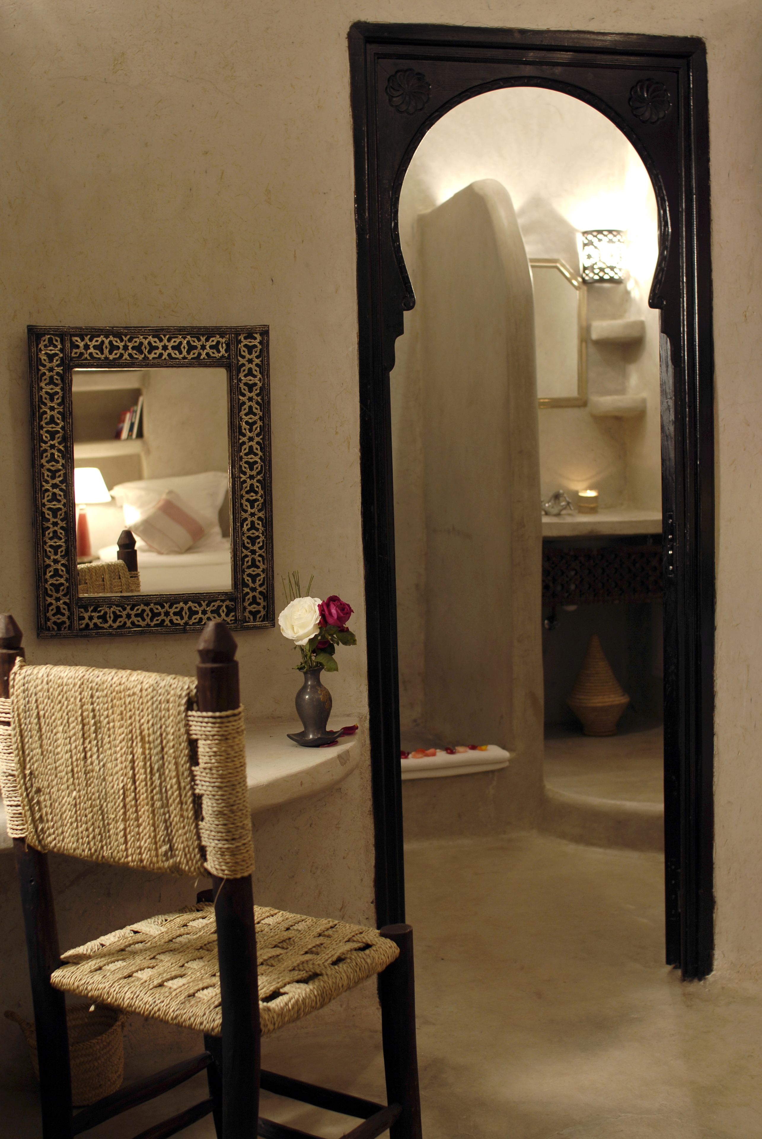 A room with a woven chair, a mirror with a decorative frame, and a vase with flowers.