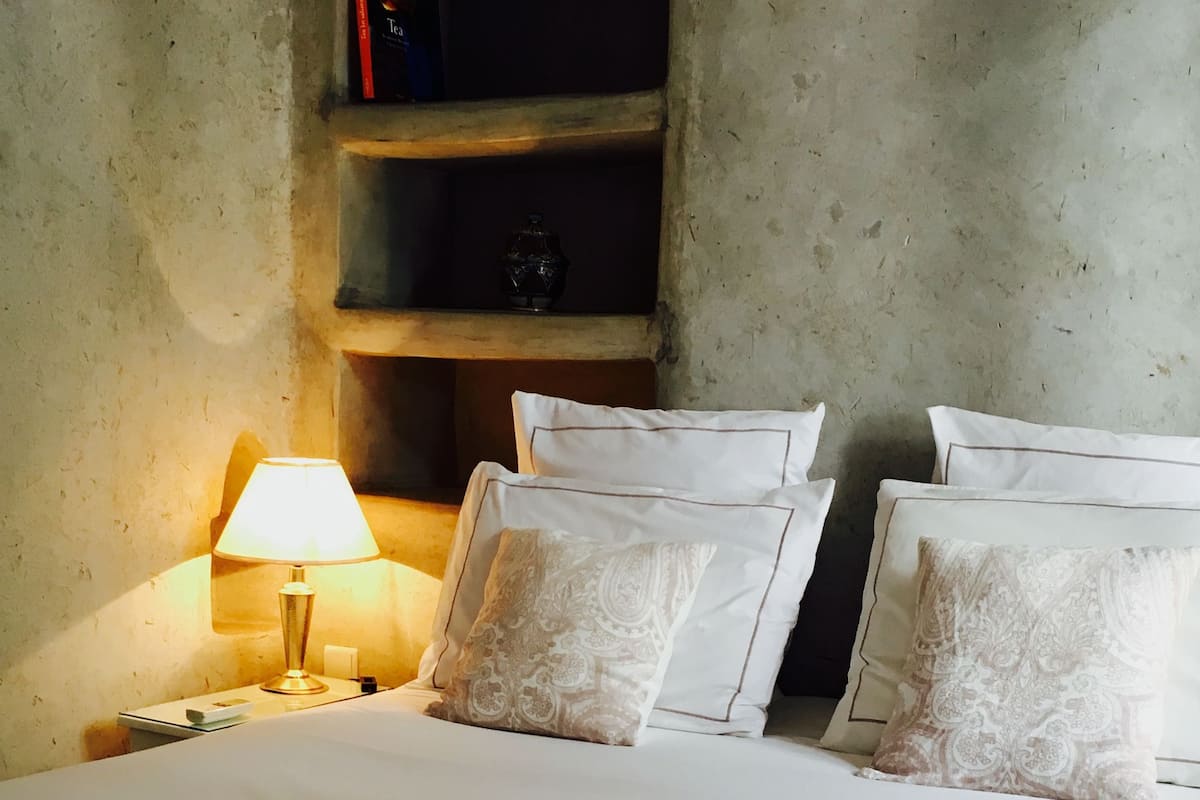 A neatly made bed with white linens and decorative pillows, a bedside lamp, and a shelf with books and a decorative item.