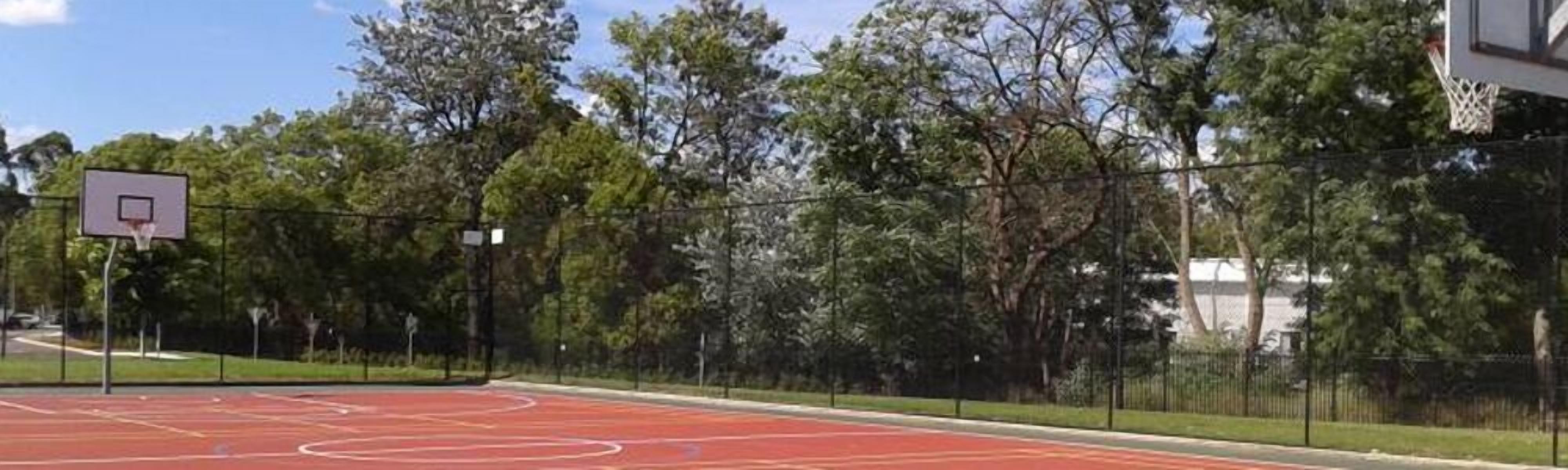 An outdoor basketball court with a red surface, surrounded by a black fence and trees.