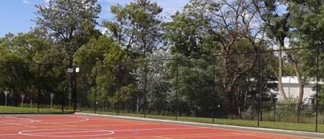 An outdoor basketball court with a red surface, surrounded by a black fence and trees.