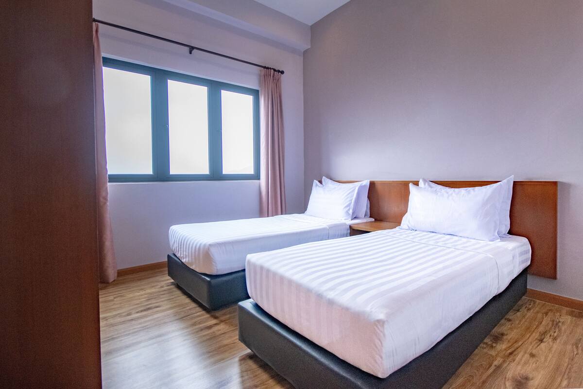 Quarto de hotel com duas camas, uma janela grande com cortinas e piso de madeira.