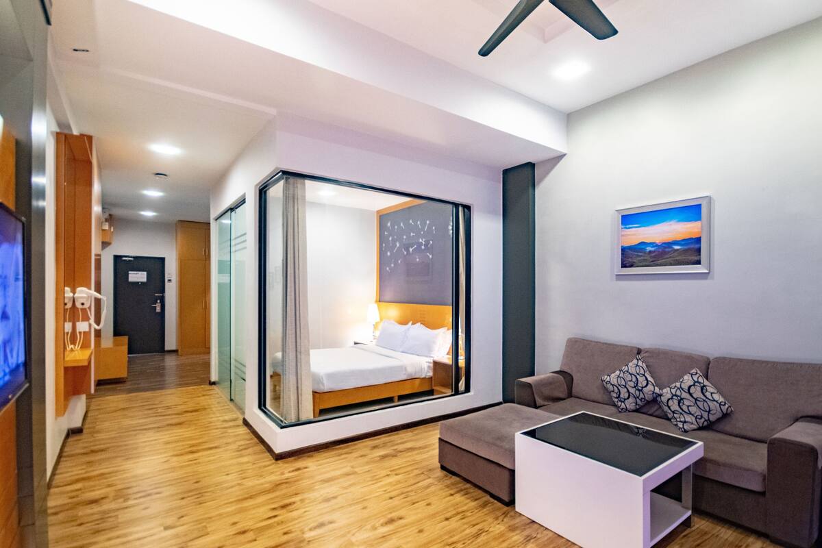 Quarto de hotel moderno com cama em estrutura de vidro, um sofá, uma mesa de centro e um quadro na parede.