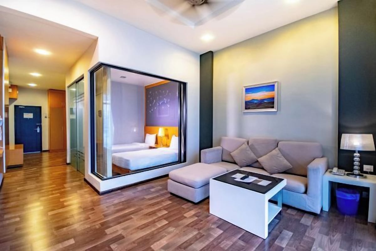 A modern hotel room with a glass partition separating the bedroom from the living area, a sofa, a coffee table, and a painting on the wall.