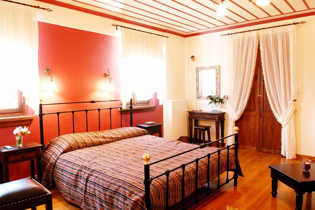 Quarto com cama, mesas de cabeceira, cômoda, espelho e porta com cortinas.