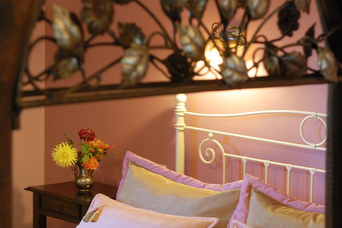 Cama com colcha amarela e várias almofadas, mesa de cabeceira com um vaso de flores e cabeceira decorativa de metal.