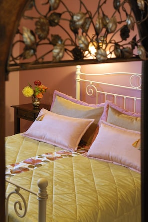 Una cama con colcha amarilla y varias almohadas, una mesita de noche con un jarrón de flores y un cabecero decorativo de metal.