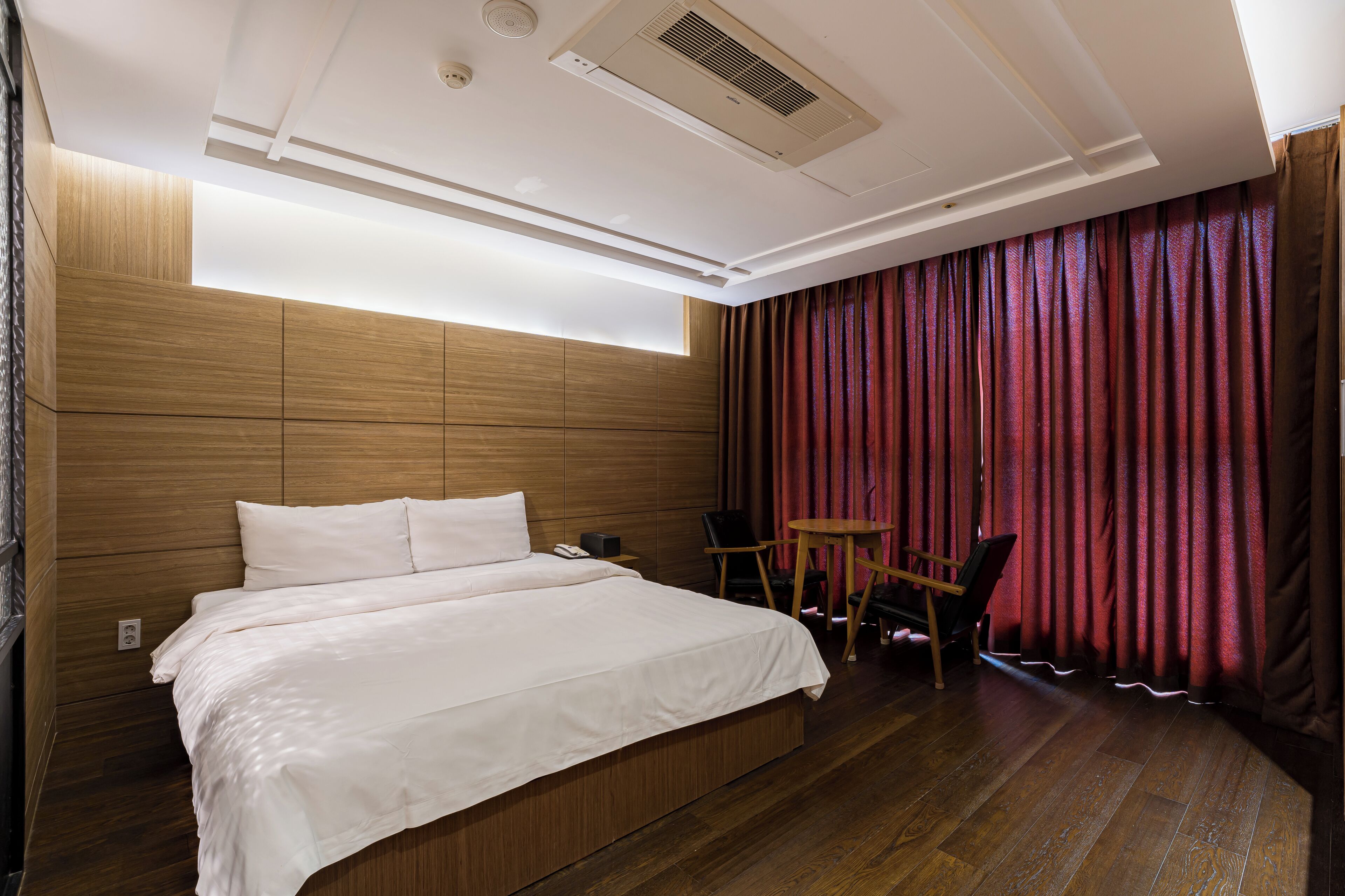 A hotel room with a large bed, wooden headboard, a desk with a chair, and a large window with curtains.