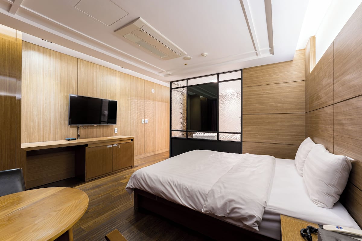 A modern hotel room with a wooden wall panel, a flat-screen TV, a bed with white linens, a small wooden table, and a desk with a chair.
