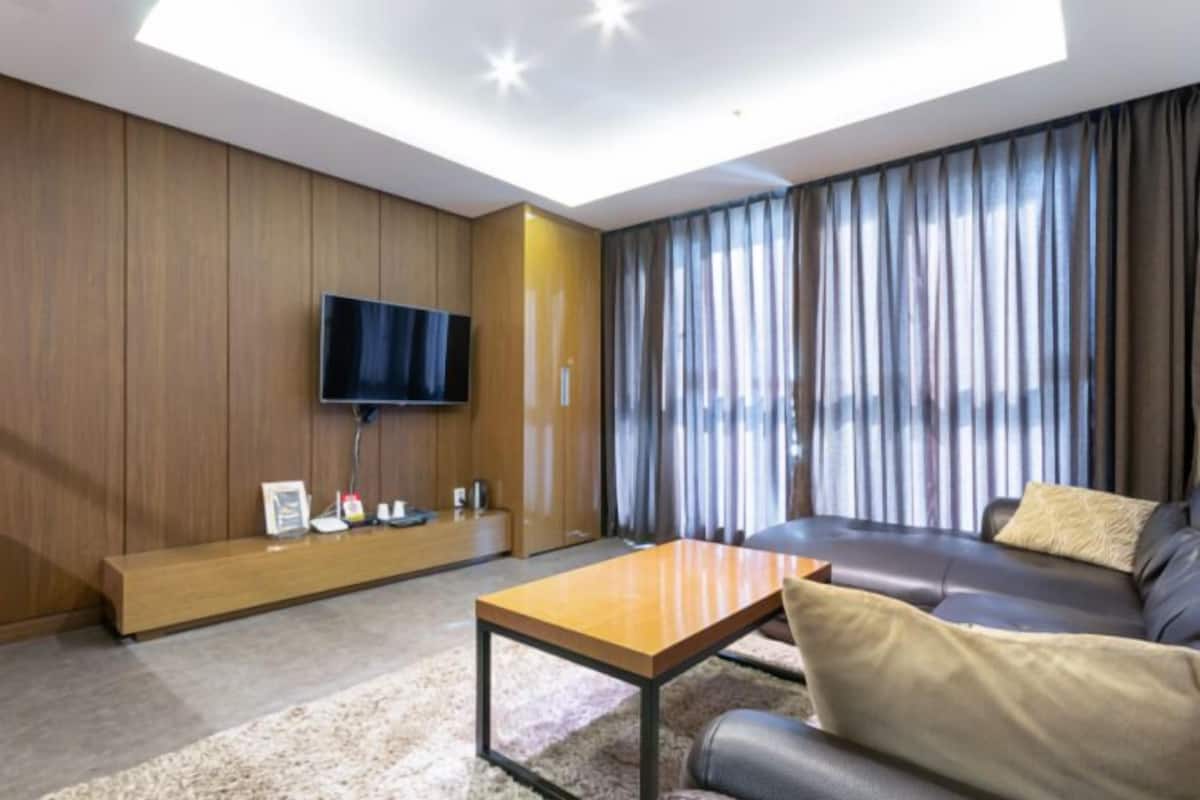 A modern living room with a flat-screen TV mounted on a wooden wall, a leather sofa, a wooden coffee table, and a large window with curtains.