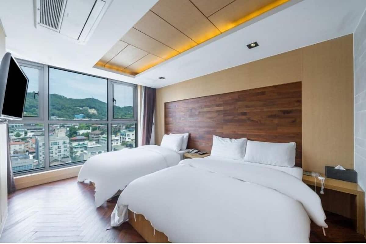 A hotel room with two beds, a wooden headboard, a large window with a city view, and a flat-screen TV.