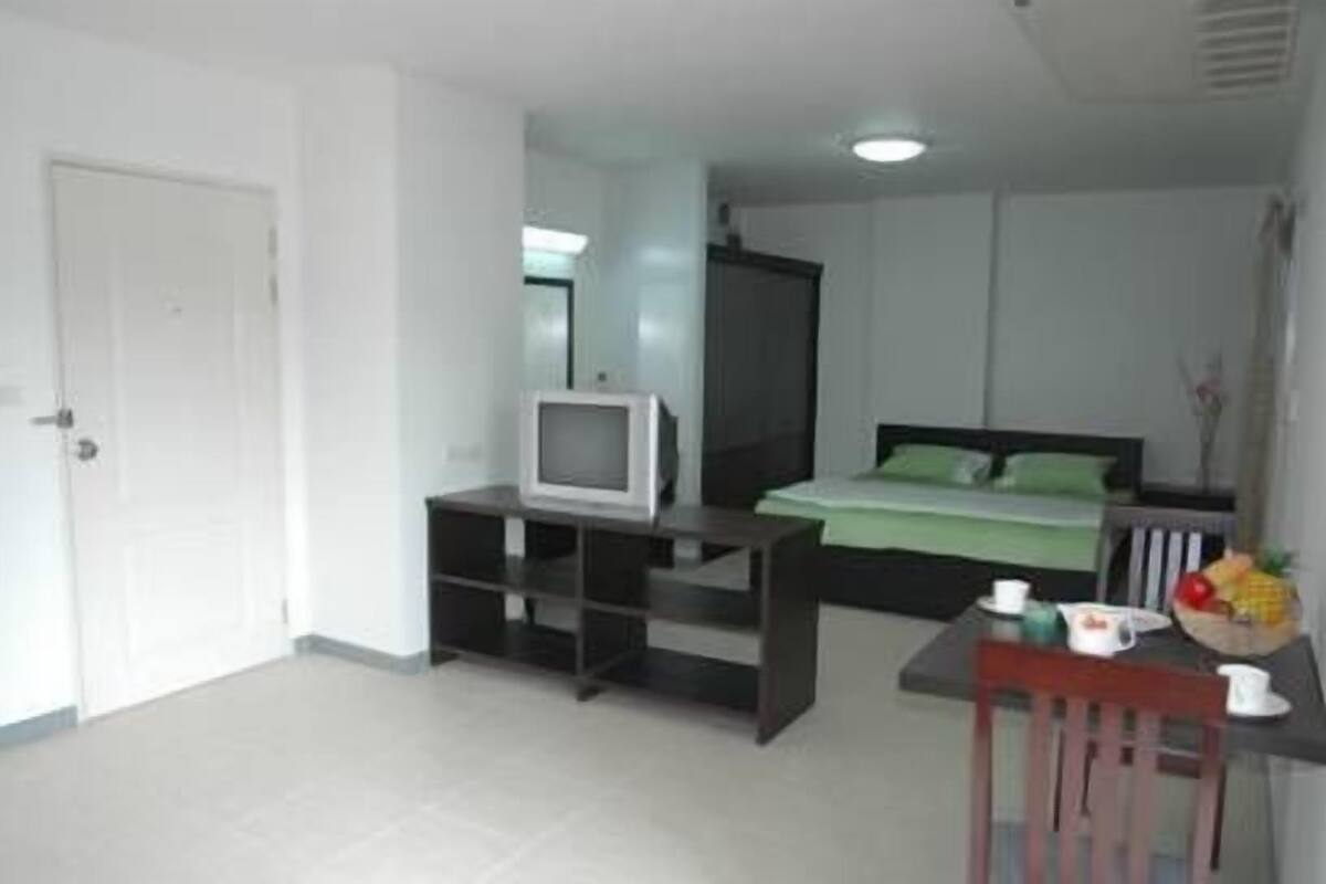 Una habitación pequeña y luminosa con una cama, un televisor sobre un mueble bajo, un escritorio con una silla y una mesita con un frutero.