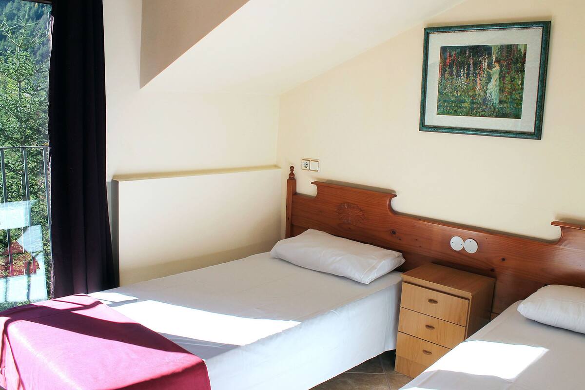 A bedroom with a single bed, a nightstand, a wooden headboard, a framed picture on the wall, and a window with a view of trees.