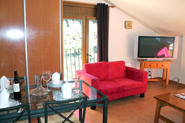 A living room with a red sofa, a glass dining table with chairs, a television, and a balcony with a view of trees.