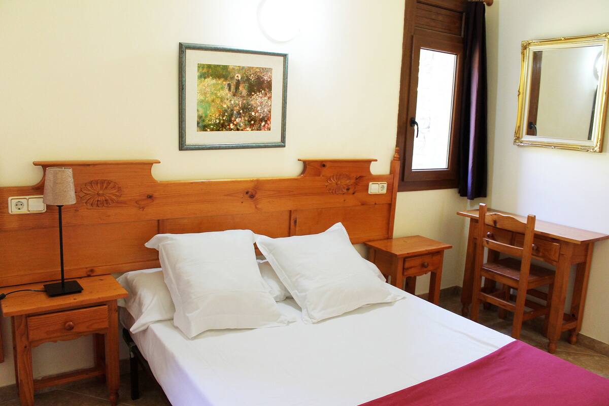 A neatly made bed with white linens and pillows, a bedside table with a lamp, a wooden nightstand, a wooden chair, and a framed picture on the wall.