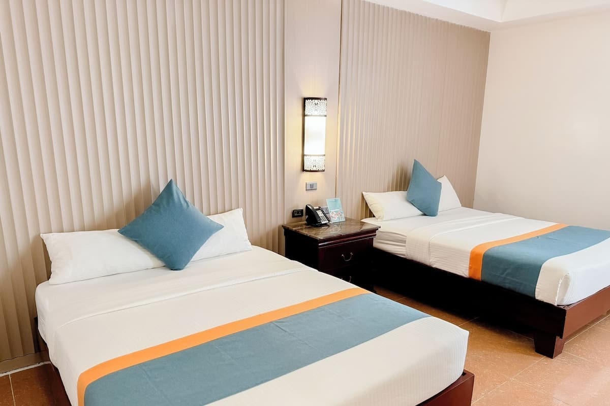 Two beds with white bedding and blue accents, a nightstand with a phone and lamp, and a wall-mounted light fixture.