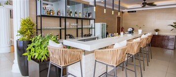 Una zona de bar con una barra larga, sillas de mimbre, una planta grande en maceta y una vitrina de vidrio con botellas.