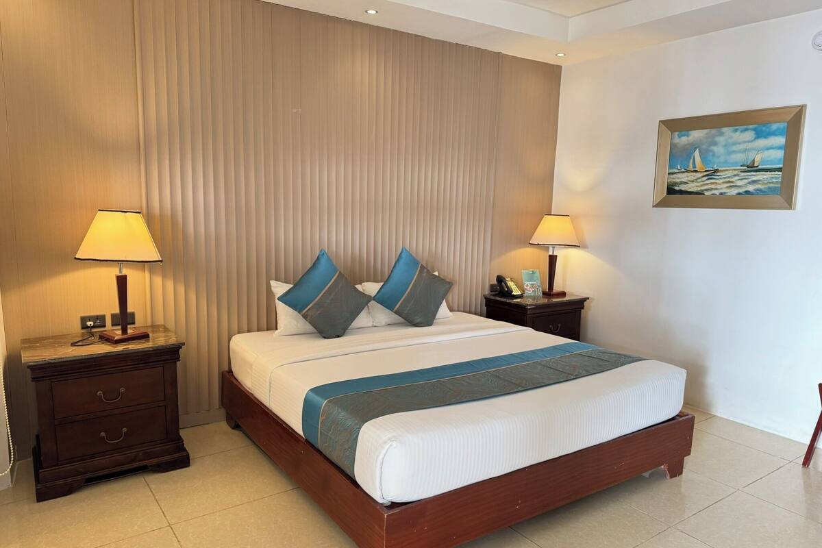 A hotel room with a large bed, two bedside tables, a nightstand, a painting on the wall, and a window with blinds.