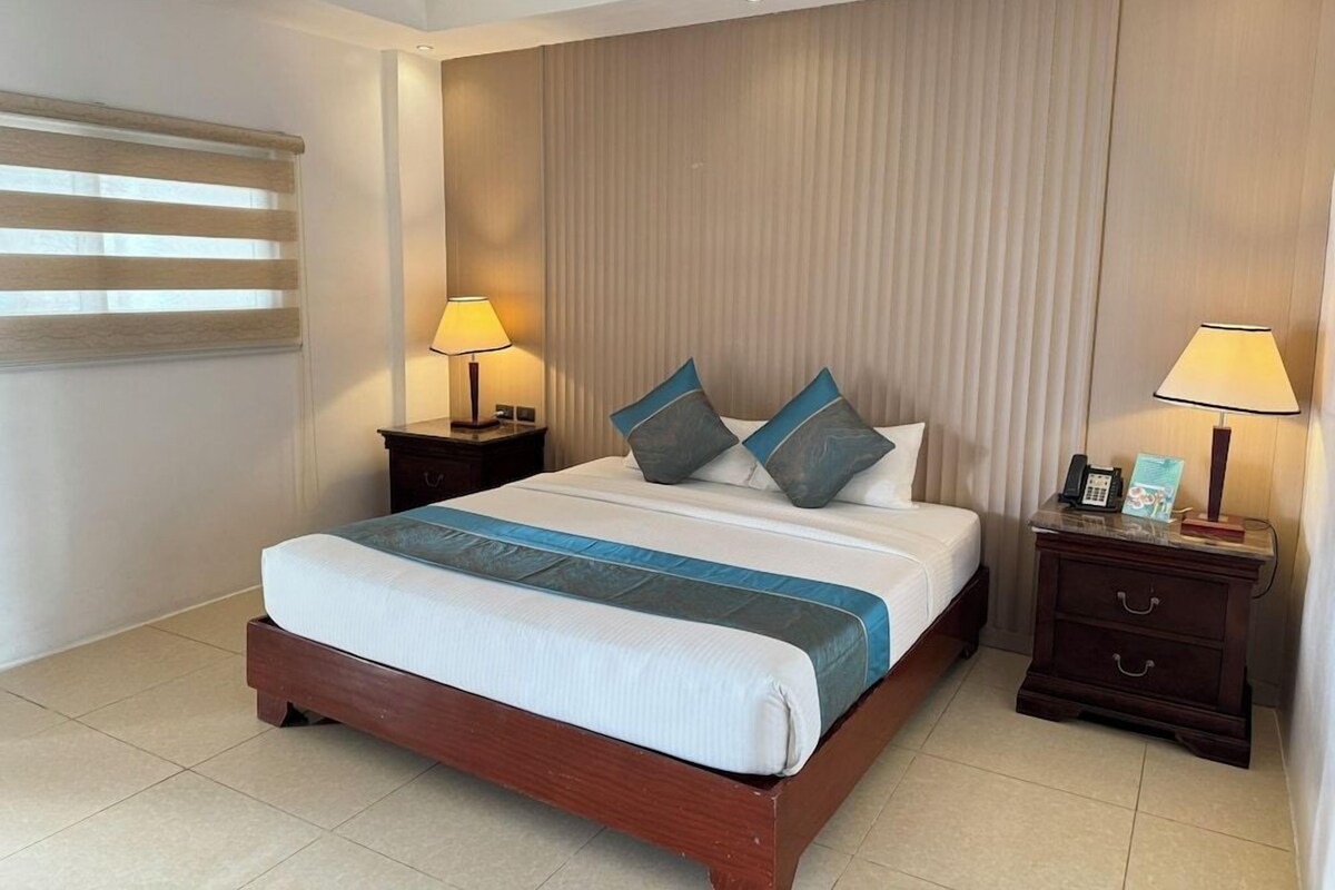 Een hotelkamer met een groot bed, twee nachtkastjes, een lamp en een raam met jaloezieën.