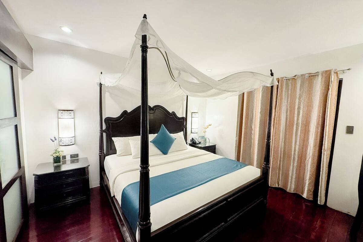 A four-poster bed with a canopy, a bedside table with a lamp, and a window with curtains.